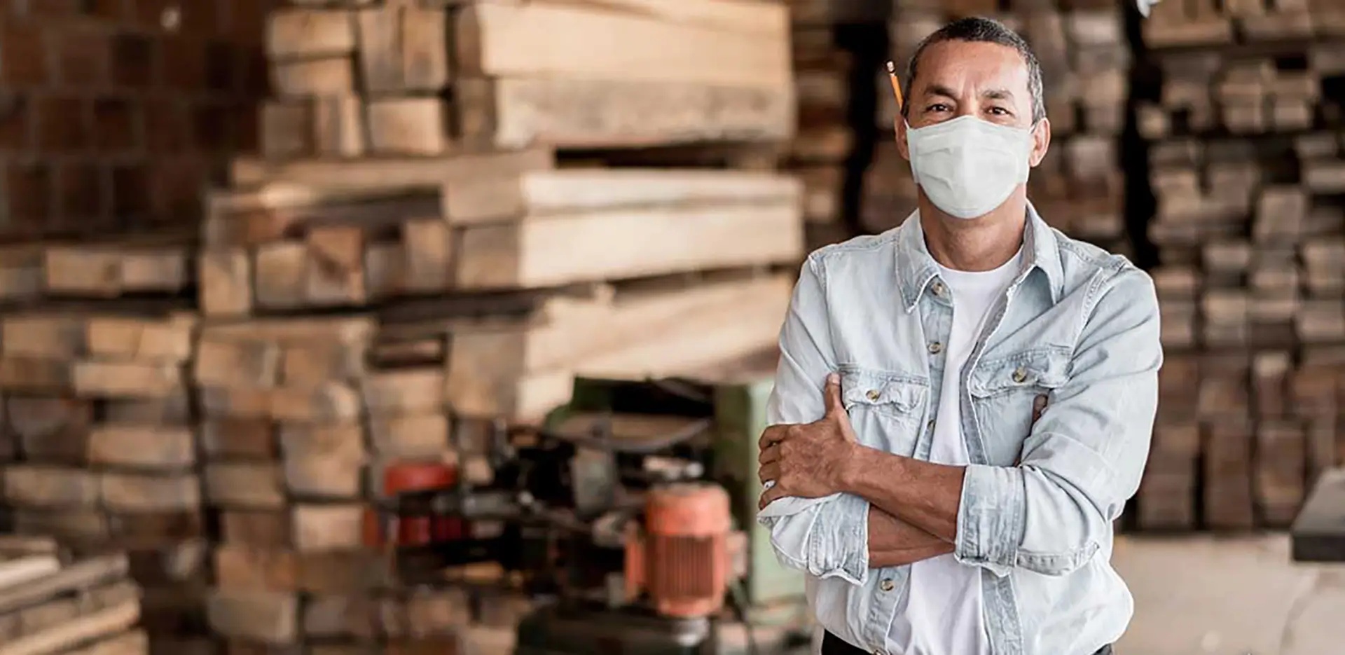 A man with a pencil over his ear wearing a mask. He's standing in front of a pile of lumber.
