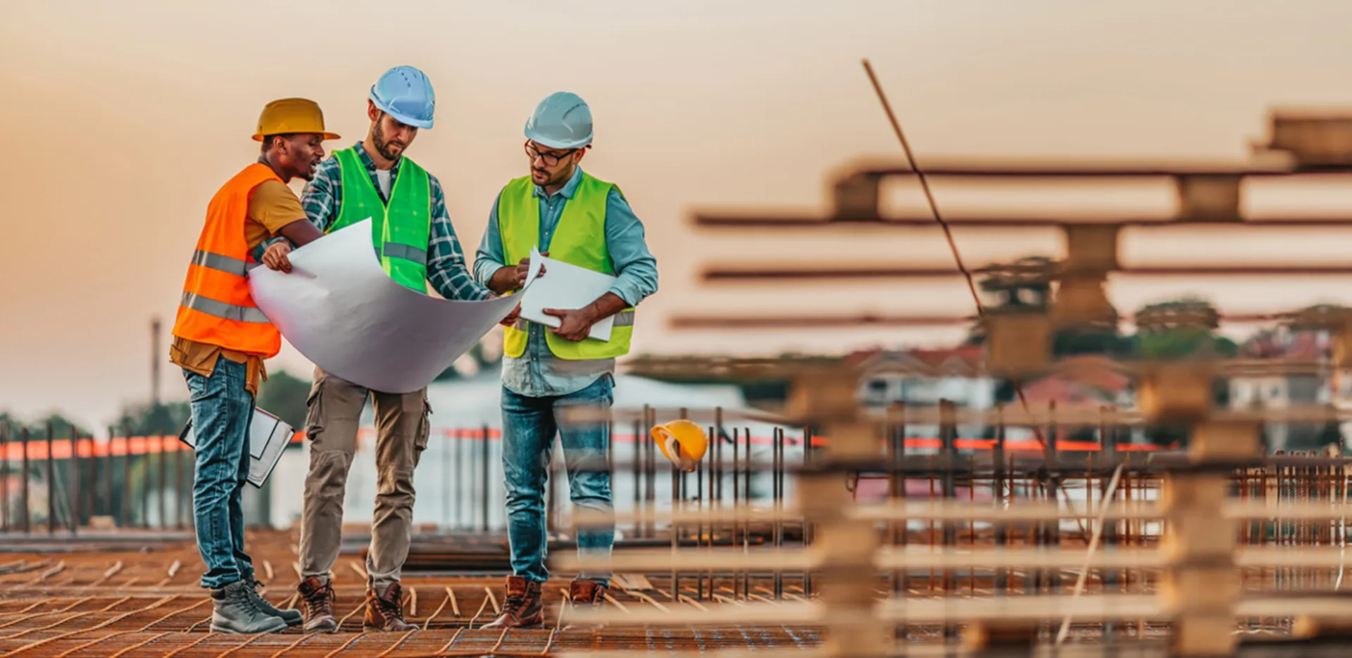 Three construction workers in safety gear review blueprints on a jobsite.