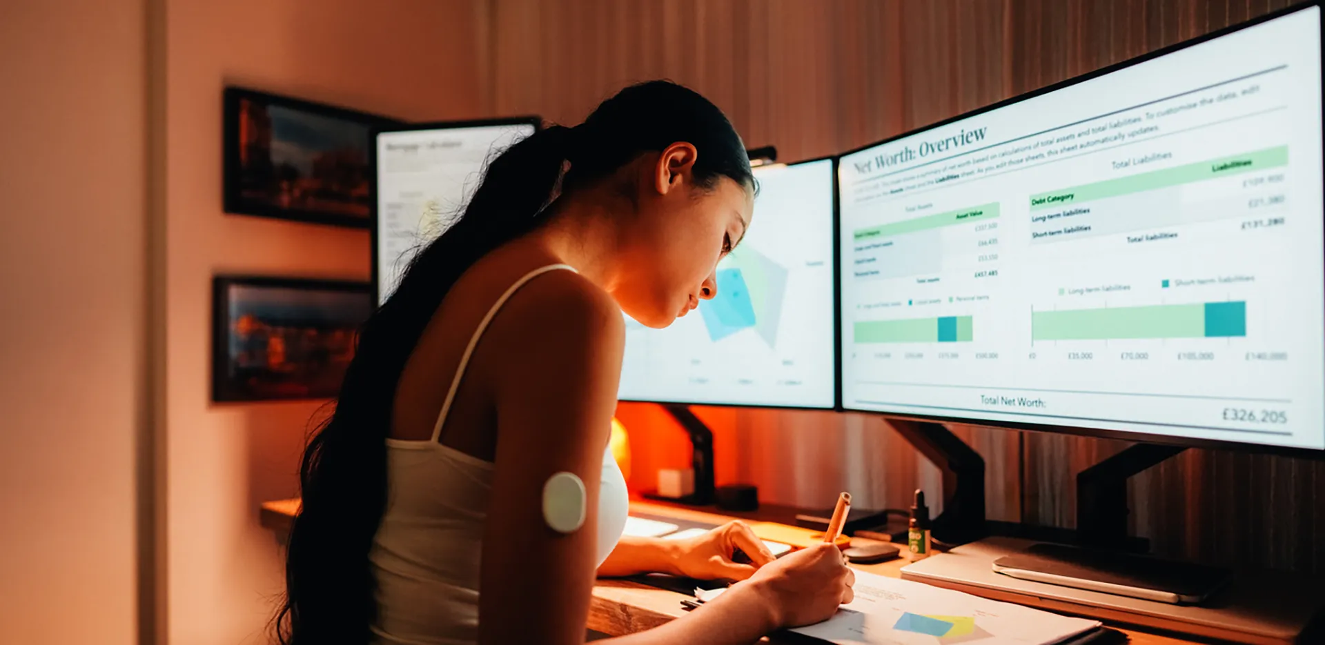 A woman taking notes while looking at graphs on a computer monitor.