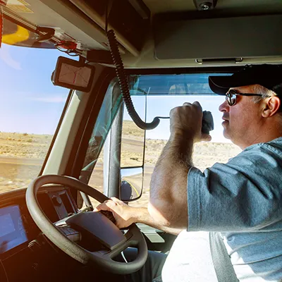 A truck driver in a truck cab, driving and talking on a CB radio.