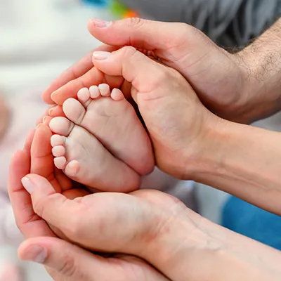 A baby's feet being cupped by two pairs of hands. An out-of-focus baby is in the background.