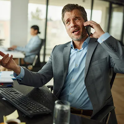 A distressed business man talking on a phone while looking at a computer monitor.