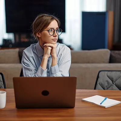A young woman sitting at a table with a laptop, staring away in the distance.