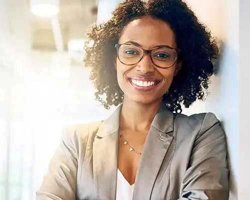 A woman wearing a grey jacket and glasses smiling as she leans against a wall.