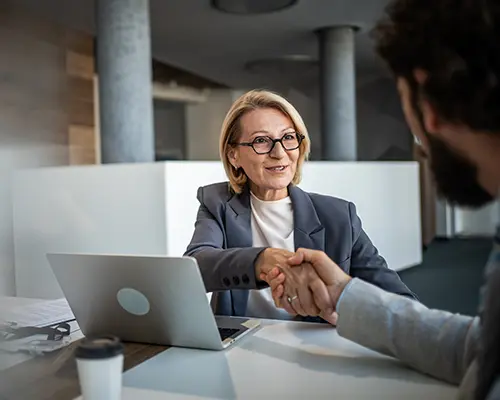 A woman with glasses sitting at a table with a laptop and shaking the hand of a man across form her.