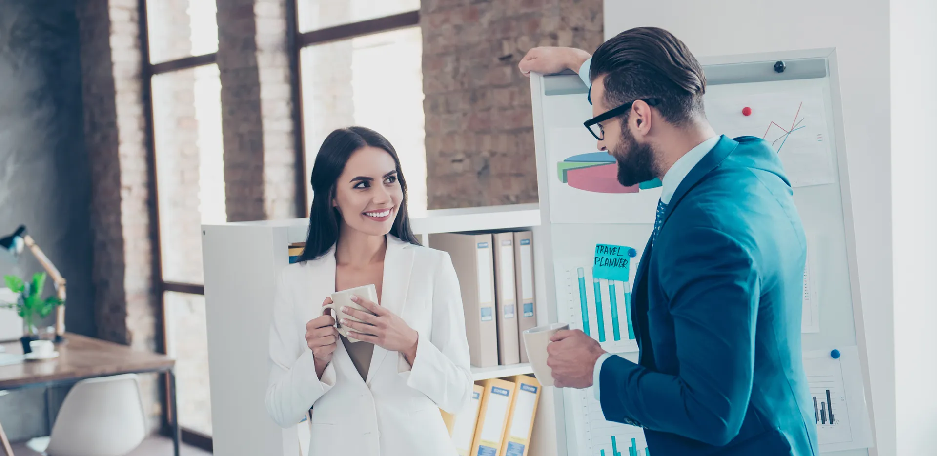 Two people dressed professionally smiling at each other while holding coffee cups.