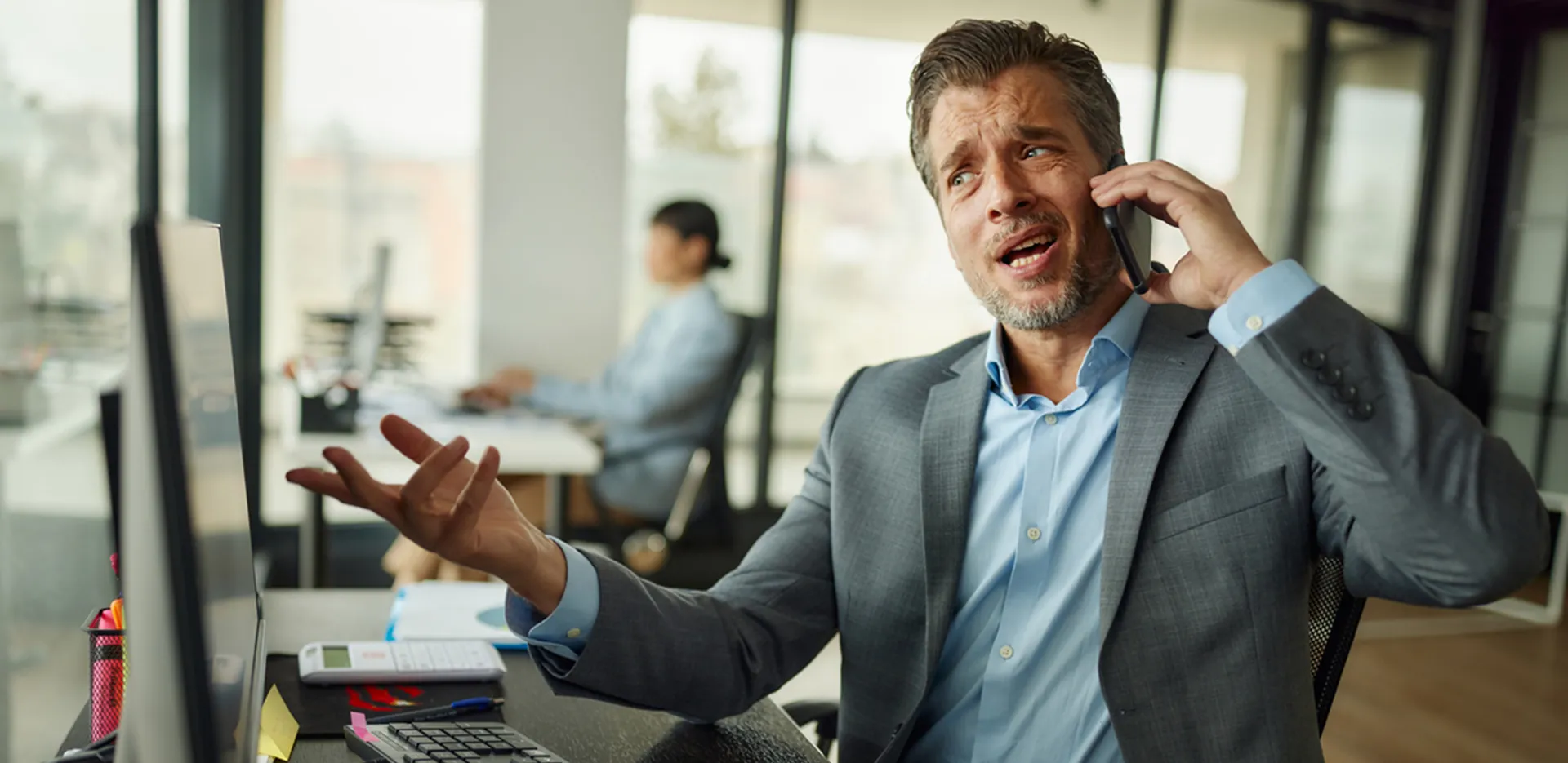 A distressed business man talking on a phone while looking at a computer monitor.