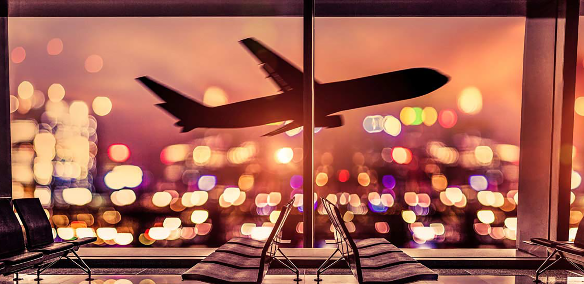 An airplane in flight seen from the window of an airport waiting area at night.