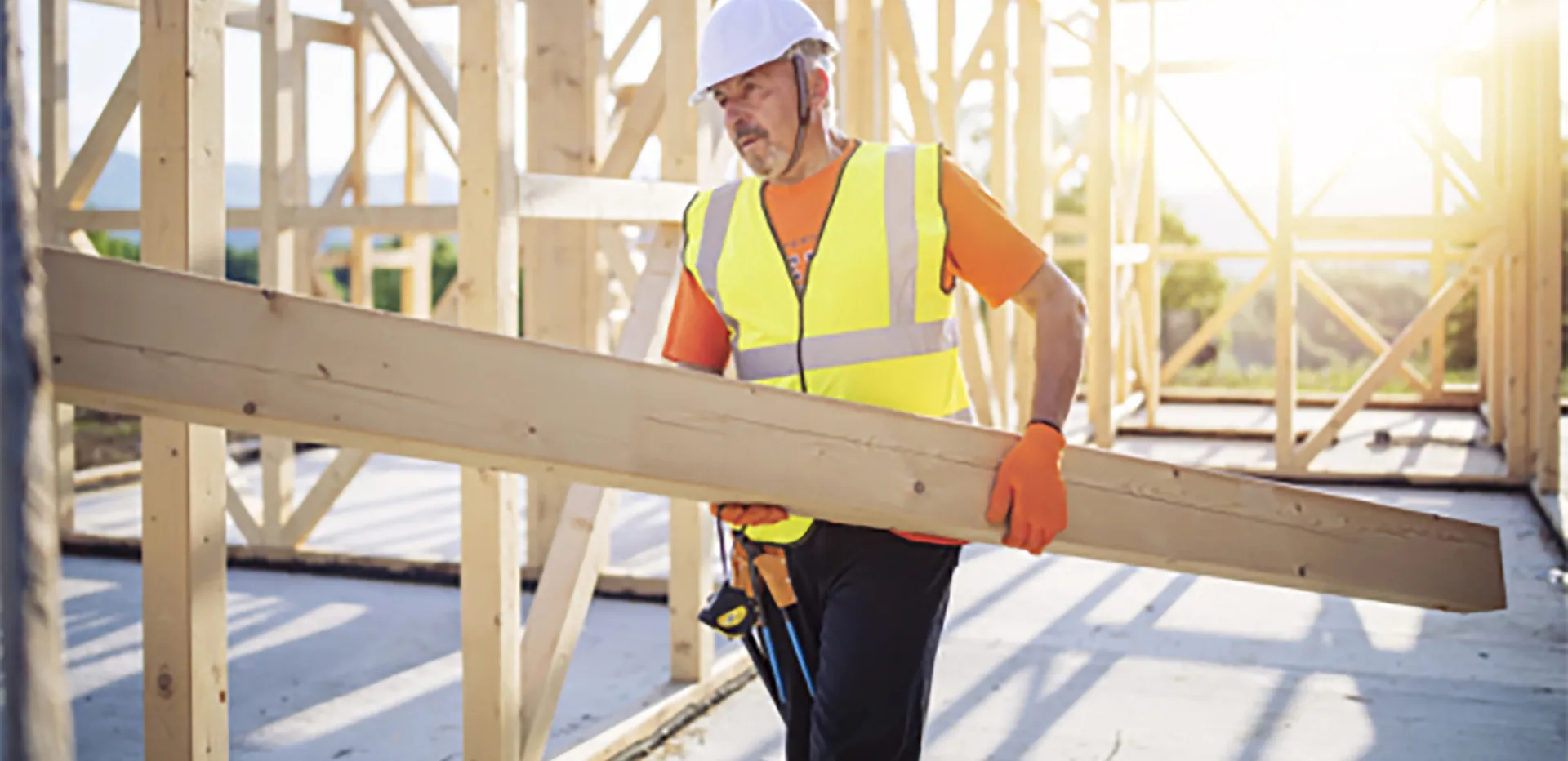 A construction worker wearing safety gear carrying a wooden beam.