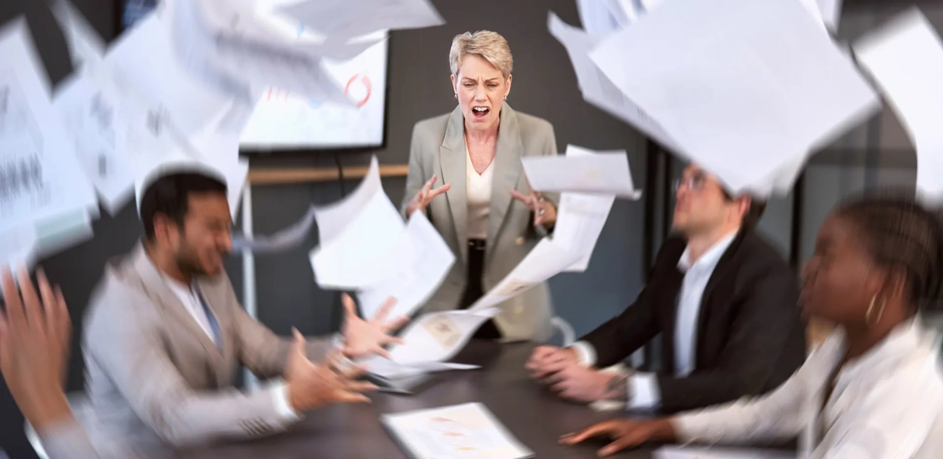 Upset business people at a conference table with papers flying through the air around them.